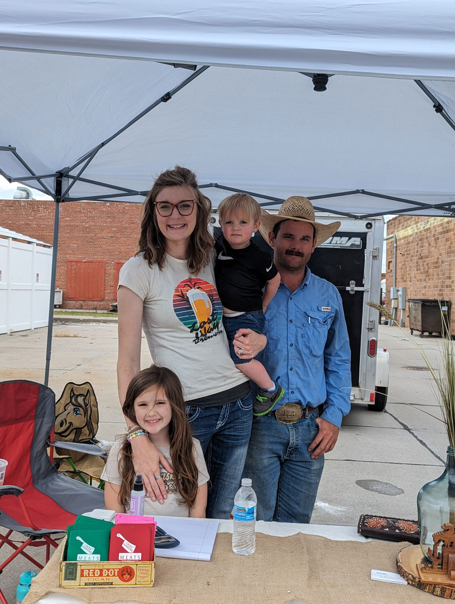 Family standing under a canopy at an outdoor event, with table displaying items in foreground.