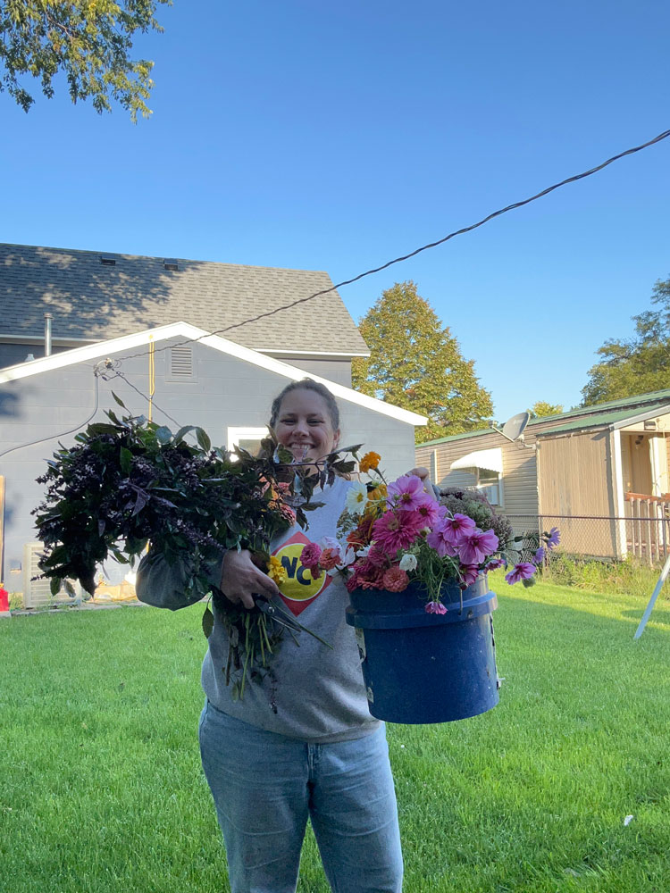 Person holding vibrant flowers and a blue bucket in a grassy backyard under a clear blue sky.